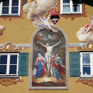 Lüftlmalerei auf einem Haus in Mittenwald mit Jesus am Kreuz in einer Passionsdarstellung. , © Alpenwelt Karwendel | Rudolf Pohmann